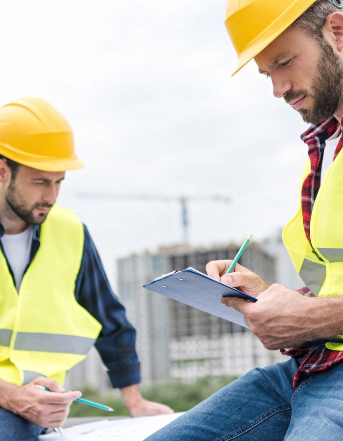 two engineers in safety vests and hardhats working with blueprints and clipboard on roof
